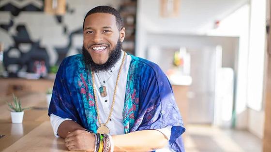 Rico Robinson stands in a café. 他穿着一件白色系扣衬衫，外面裹着一件蓝色图案的围巾. He is smiling.